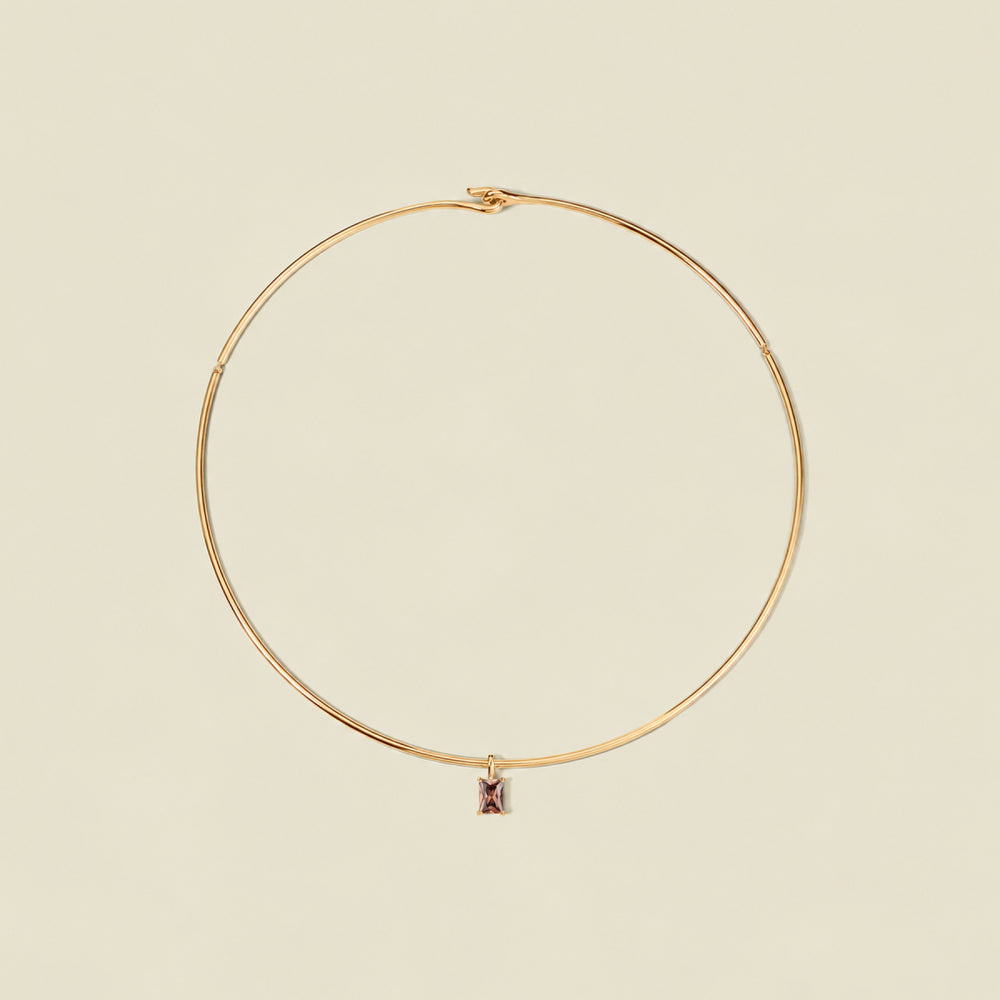 A thin gold wire hoop necklace with a small rectangular black-purple baguette gemstone charm, displayed on a solid light beige background.