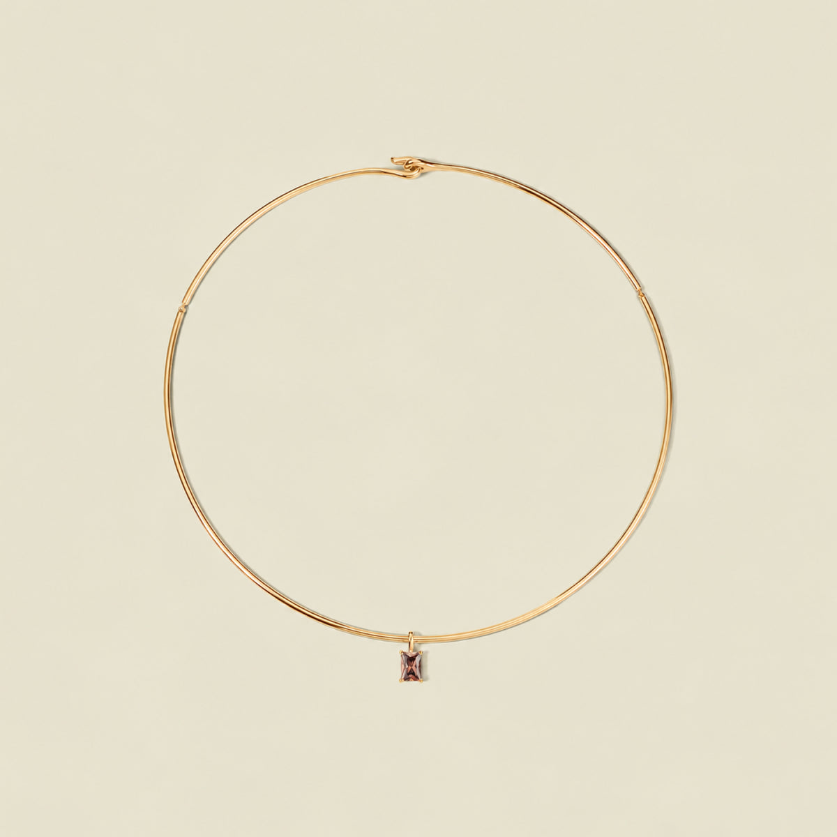 A thin gold wire hoop necklace with a small rectangular black-purple baguette gemstone charm, displayed on a solid light beige background.