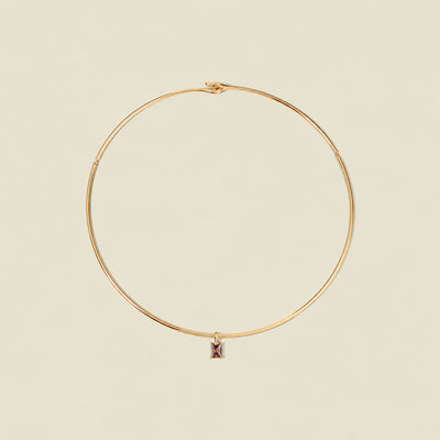 A thin gold wire hoop necklace with a small rectangular black-purple baguette gemstone charm, displayed on a solid light beige background.