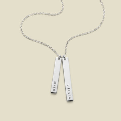 A silver double-bar necklace with personalized names engraved on each bar, hanging vertically from a delicate silver chain against a solid light beige background.