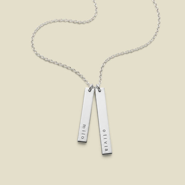 A silver double-bar necklace with personalized names engraved on each bar, hanging vertically from a delicate silver chain against a solid light beige background.