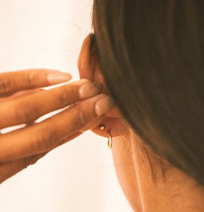 A close-up lifestyle shot of the back of a person's ear wearing a small gold hoop earring, with their hand adjusting the earring on their ear. The background is a soft, warm off-white.