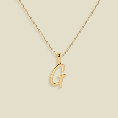 A gold initial necklace featuring a polished cursive letter 'G' pendant on a delicate gold chain, set against a solid light beige background.