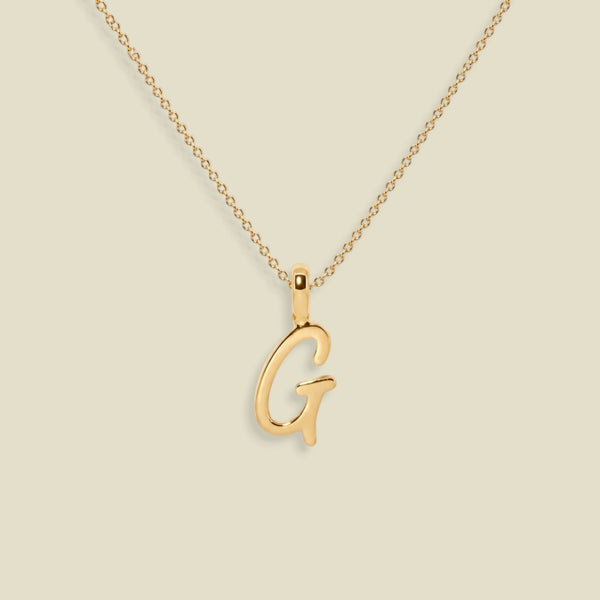 A gold initial necklace featuring a polished cursive letter 'G' pendant on a delicate gold chain, set against a solid light beige background.