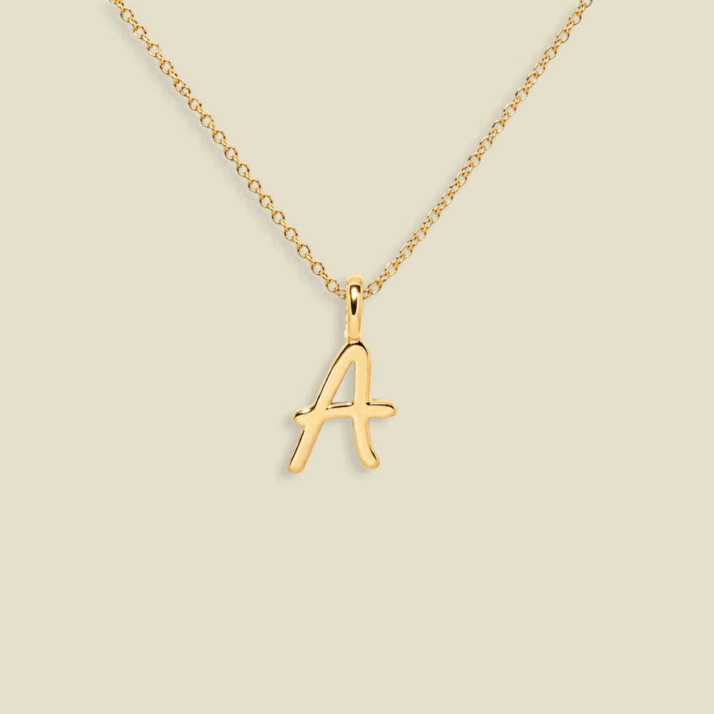 A gold initial necklace featuring a cursive, openwork letter 'A' pendant on a delicate gold chain, set against a solid light beige background.