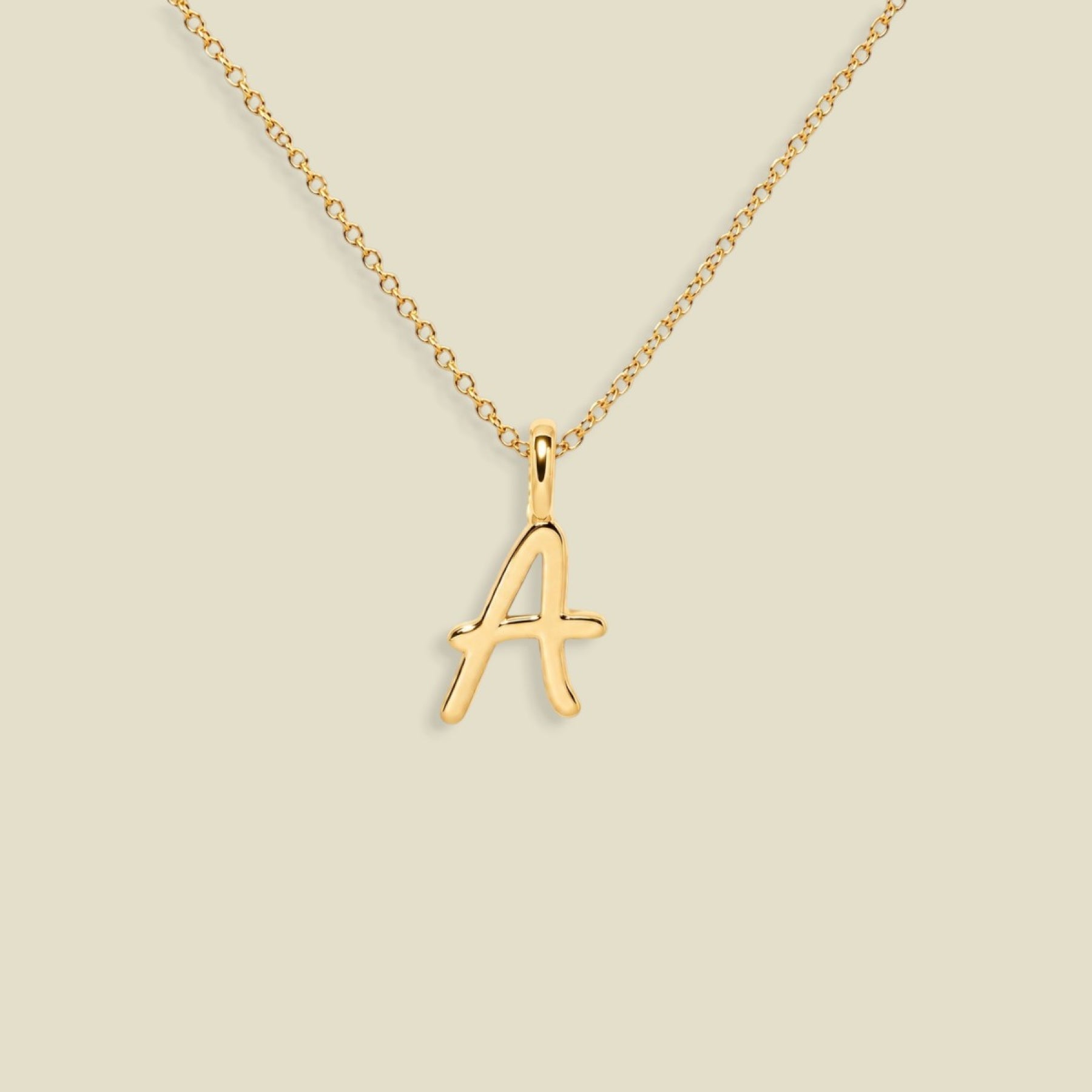 A gold initial necklace featuring a cursive, openwork letter 'A' pendant on a delicate gold chain, set against a solid light beige background.