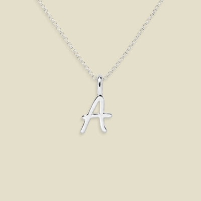 A silver initial necklace featuring a polished cursive letter 'A' pendant on a delicate silver chain, set against a solid light beige background.