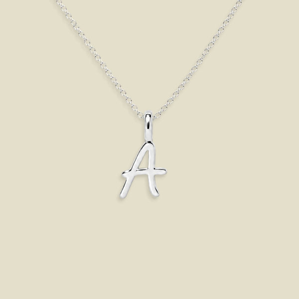 A silver initial necklace featuring a polished cursive letter 'A' pendant on a delicate silver chain, set against a solid light beige background.