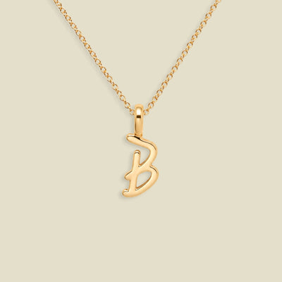 A gold initial necklace featuring a polished cursive letter 'B' pendant on a delicate gold chain, set against a solid light beige background.