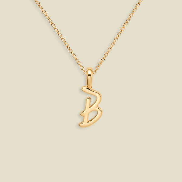 A gold initial necklace featuring a polished cursive letter 'B' pendant on a delicate gold chain, set against a solid light beige background.