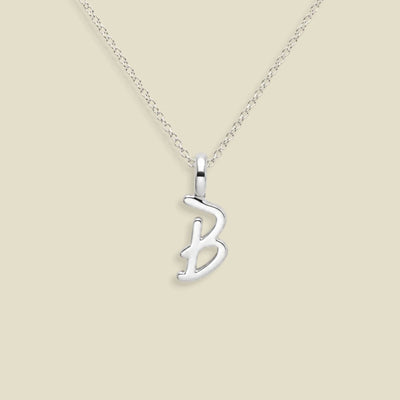 A silver initial necklace featuring a polished cursive letter 'B' pendant on a delicate silver chain, set against a solid light beige background.