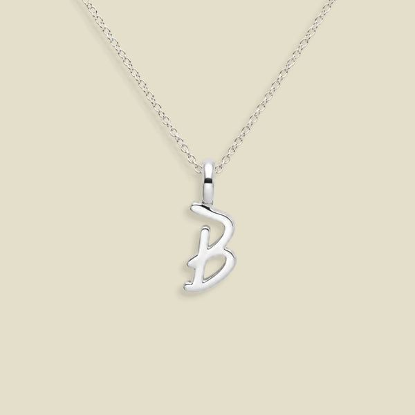 A silver initial necklace featuring a polished cursive letter 'B' pendant on a delicate silver chain, set against a solid light beige background.