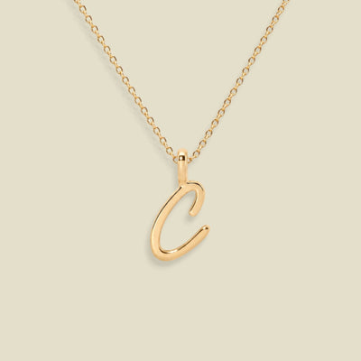 A gold initial necklace featuring a polished cursive letter 'C' pendant on a delicate gold chain, set against a solid light beige background.