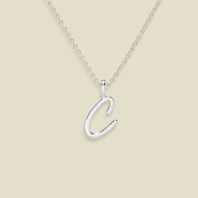 A silver initial necklace featuring a polished cursive letter 'C' pendant on a delicate silver chain, set against a solid light beige background.