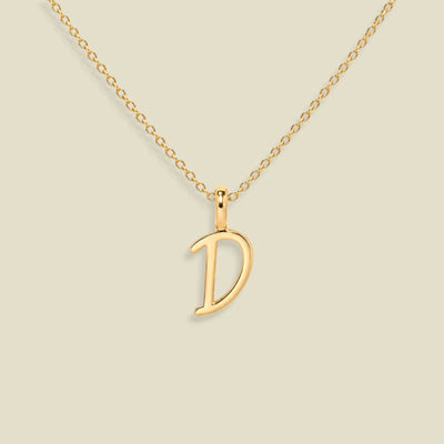 A gold initial necklace featuring a polished open letter script font 'D' pendant on a delicate gold chain, set against a solid light beige background.