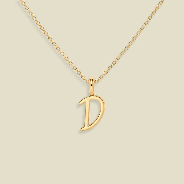 A gold initial necklace featuring a polished open letter script font 'D' pendant on a delicate gold chain, set against a solid light beige background.