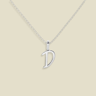 A silver initial necklace featuring a polished script font 'D' pendant on a delicate cable chain, set against a solid light beige background.