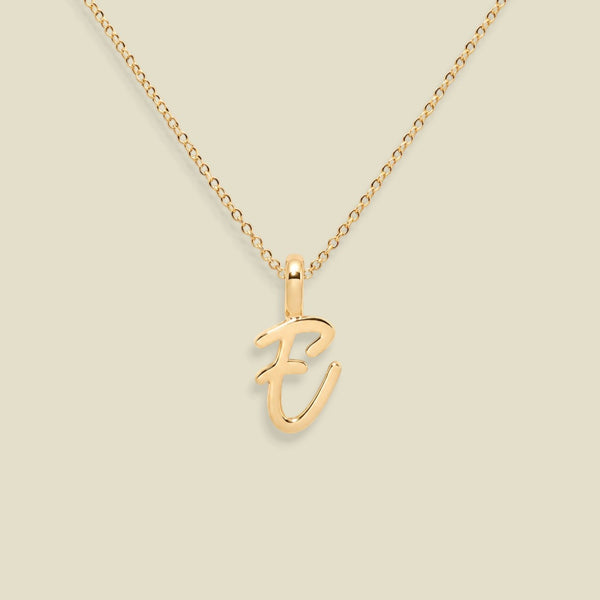 A gold initial pendant necklace featuring the letter 'E' in a cursive script, suspended from a delicate gold cable chain against a solid light beige background.
