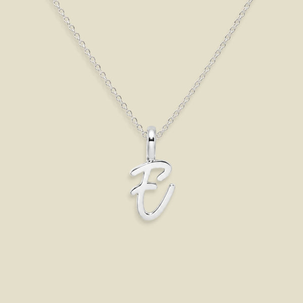 A silver initial necklace featuring a polished cursive letter 'E' pendant on a delicate cable chain, set against a solid light beige background.