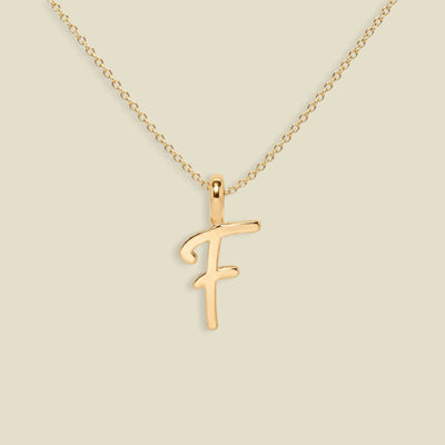 A gold initial necklace featuring a cursive script letter 'F' pendant on a delicate gold chain, set against a solid light beige background.