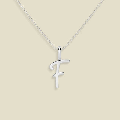 A silver initial pendant necklace featuring the letter 'F' in a cursive script, suspended from a delicate silver chain against a solid beige background.