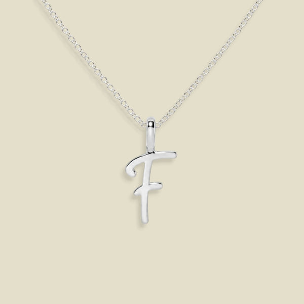 A silver initial pendant necklace featuring the letter 'F' in a cursive script, suspended from a delicate silver chain against a solid beige background.