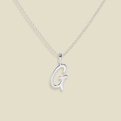 A silver initial necklace featuring a polished cursive letter 'G' pendant on a delicate silver chain, set against a solid light beige background.