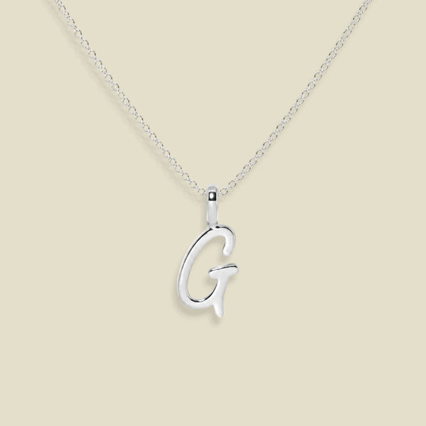 A silver initial necklace featuring a polished cursive letter 'G' pendant on a delicate silver chain, set against a solid light beige background.