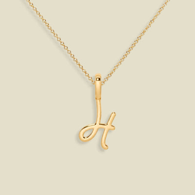 A gold initial pendant necklace featuring the letter 'H' in a polished script design, suspended from a delicate gold chain against a solid light beige background.