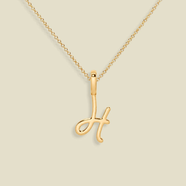 A gold initial pendant necklace featuring the letter 'H' in a polished script design, suspended from a delicate gold chain against a solid light beige background.
