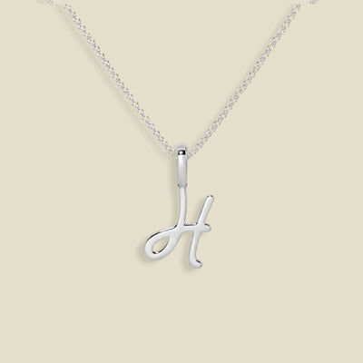 A silver initial necklace featuring a polished cursive letter 'H' pendant on a delicate silver chain, set against a solid light beige background.