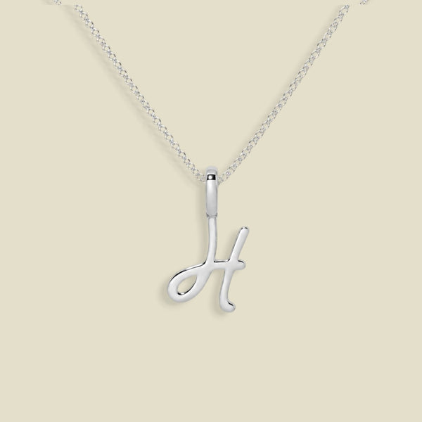 A silver initial necklace featuring a polished cursive letter 'H' pendant on a delicate silver chain, set against a solid light beige background.
