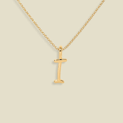 A gold initial necklace featuring a polished letter 'I' pendant on a delicate gold chain, set against a solid light beige background.