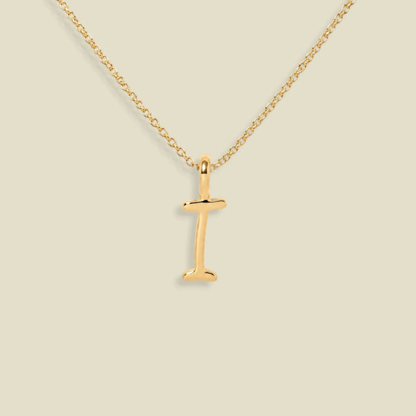 A gold initial necklace featuring a polished letter 'I' pendant on a delicate gold chain, set against a solid light beige background.