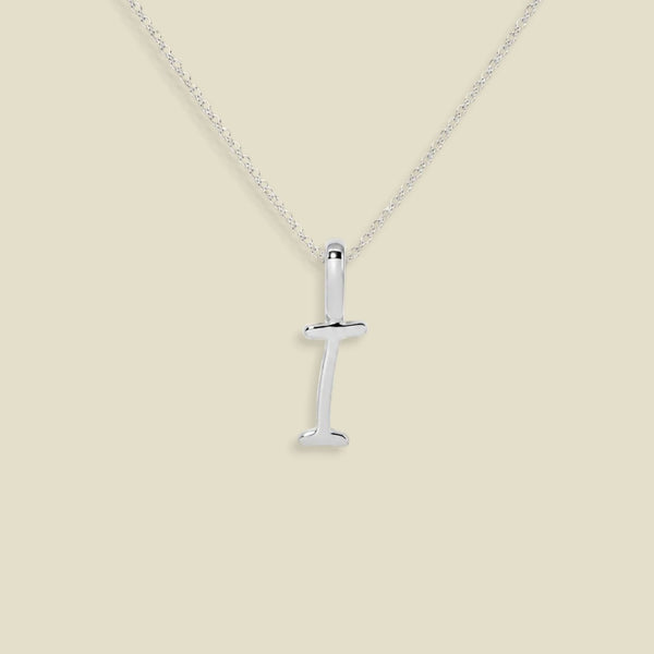 A silver initial necklace featuring a polished letter 'I' pendant in script font on a delicate silver chain, set against a solid light beige background.