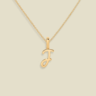 A gold initial necklace featuring a polished cursive letter 'J' pendant on a delicate gold chain, set against a solid light beige background.