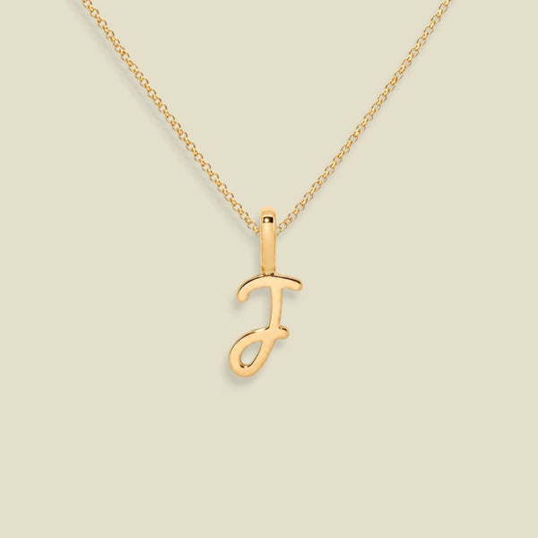 A gold initial necklace featuring a polished cursive letter 'J' pendant on a delicate gold chain, set against a solid light beige background.