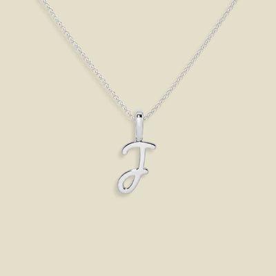 A silver initial pendant necklace featuring the uppercase letter 'J' in a cursive script, suspended from a delicate silver chain against a solid beige background.
