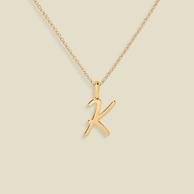 A gold initial necklace featuring a polished cursive uppercase letter 'K' pendant on a delicate gold chain, set against a solid light beige background.