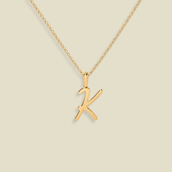 A gold initial necklace featuring a polished cursive uppercase letter 'K' pendant on a delicate gold chain, set against a solid light beige background.