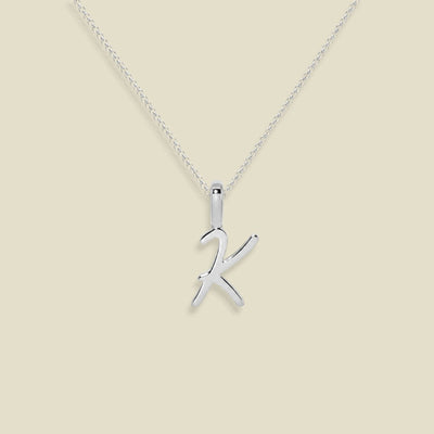 A dainty silver necklace with a polished cursive uppercase letter 'K' pendant, displayed against a solid light beige background.