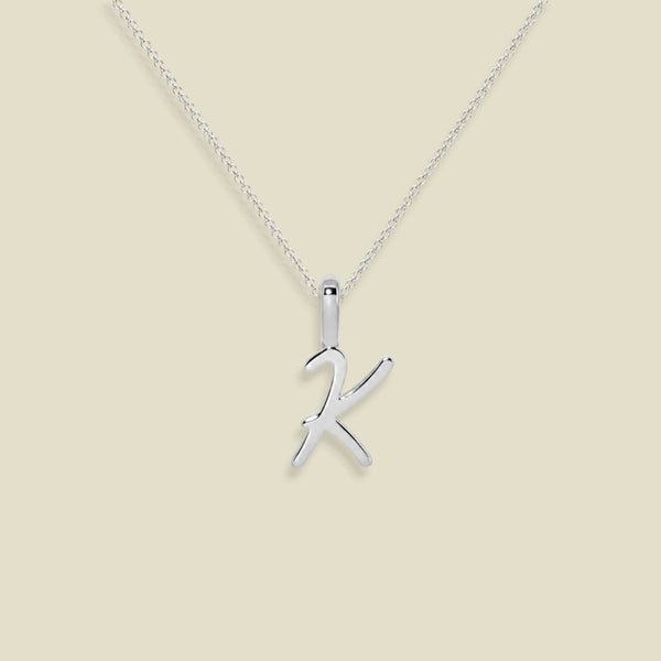 A dainty silver necklace with a polished cursive uppercase letter 'K' pendant, displayed against a solid light beige background.
