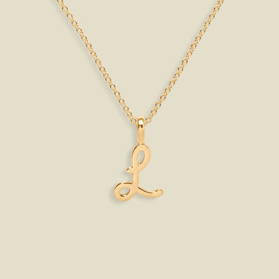 A dainty gold necklace with a polished gold cursive uppercase letter 'L' pendant, displayed against a solid light beige background.