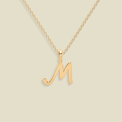 A gold initial pendant necklace featuring a polished cursive letter 'M' pendant on a delicate gold chain, set against a solid light beige background.