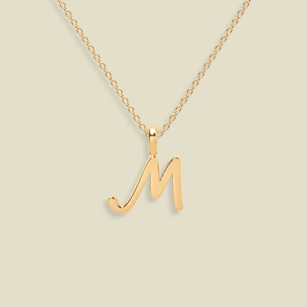 A gold initial pendant necklace featuring a polished cursive letter 'M' pendant on a delicate gold chain, set against a solid light beige background.