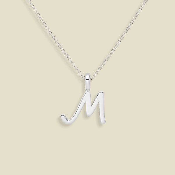 A silver initial pendant necklace featuring a polished cursive uppercase letter 'M' pendant on a delicate link chain, set against a solid light beige background.