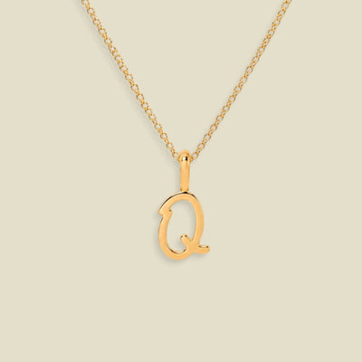 A gold initial necklace featuring a polished script uppercase letter 'Q' pendant on a delicate gold chain, set against a solid light beige background.