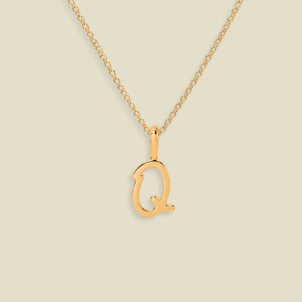 A gold initial necklace featuring a polished script uppercase letter 'Q' pendant on a delicate gold chain, set against a solid light beige background.