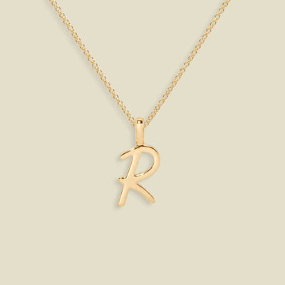 A gold initial necklace featuring a cursive uppercase-style letter 'R' pendant on a delicate gold chain, set against a solid light beige background.