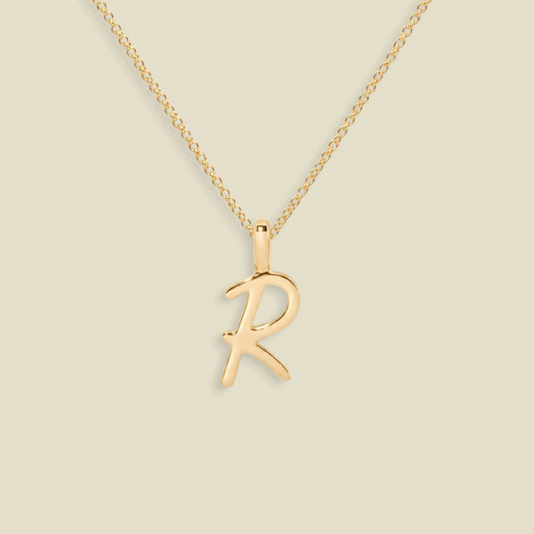 A gold initial necklace featuring a cursive uppercase-style letter 'R' pendant on a delicate gold chain, set against a solid light beige background.